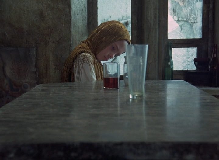 A filha do Stalker (Natasha Abramova), com um lenço dourado na cabeça, olha fixamente para os copos sobre a mesa, em Stalker (1979), de Andrei Tarkóvski.