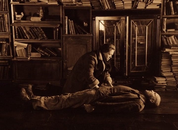 Em sépia, a mulher do Stalker (Alisa Freindlich) ajoelhada ao lado do marido (Aleksandr Kaidanóvski), deitado no chão entre estantes de livros, em Stalker (1979), de Andrei Tarkóvski.