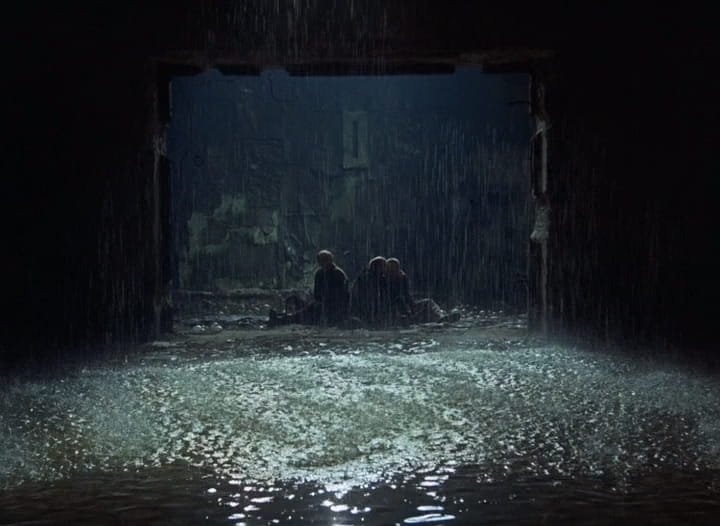 Plano geral do limiar da sala secreta: sob a chuva, o Stalker (Aleksandr Kaidanóvski), o Escritor (Anatoli Solonítsin) e o Professor (Nikolai Grinko) sentados na entrada, em Stalker (1979), de Andrei Tarkóvski.