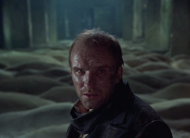O Escritor (Anatoli Solonítsin), com o rosto suado, diante de uma sala coberta por dunas de areia, em Stalker (1979), de Andrei Tarkóvski.